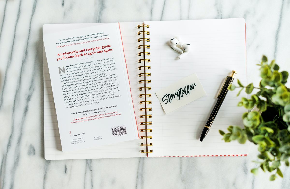 A flatlay of a workspace, with a white marble background. Includes a book, notebook, pen, earbuds and a sticker that reads "Storyteller."