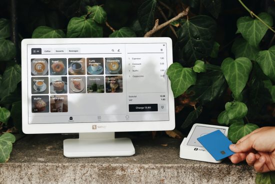 A SumUp POS system is set up in an outdoor, nature-inspired setting, with a user making a contactless payment using a blue card. The screen displays a cafe menu with items like coffee, croissants, and desserts, creating a vibrant and inviting retail experience. The clean design of the SumUp system, combined with the lush greenery surrounding it, enhances the natural, eco-friendly aesthetic of this modern payment solution, ideal for cafes, coffee shops, and small businesses.
